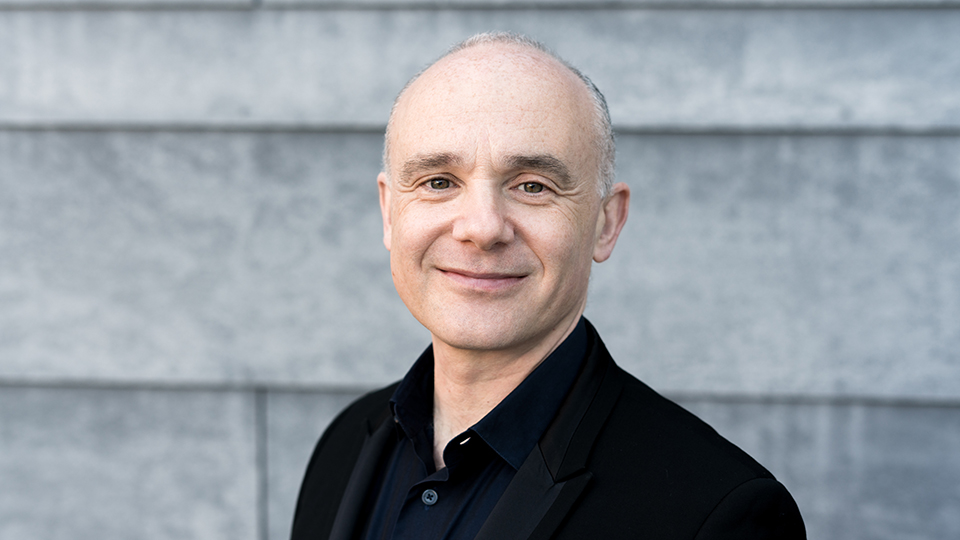 Andrew Zolinsky: A man with a dark shirt, smiling at the camera, with a grey brick building behind him.